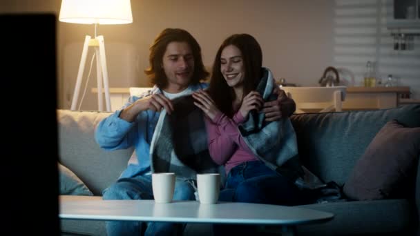 Joven chico cariñoso cubriendo a su novia con cuadros, dándole una taza de té caliente, disfrutando de una noche acogedora juntos en casa — Vídeos de Stock