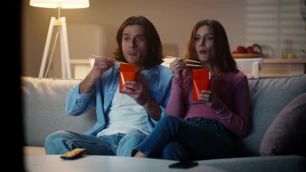 Felice giovane coppia godendo di tagliatelle asiatiche da scatole, trascorrere il fine settimana sera insieme a casa e guardare la tv — Video Stock