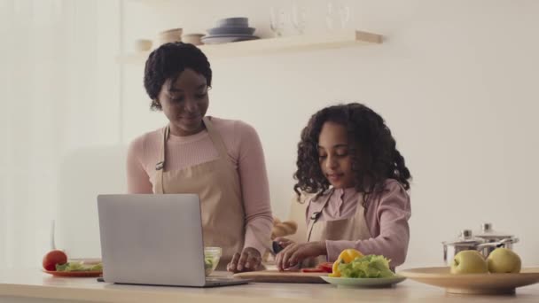 Vrolijk Afrikaans Amerikaans moeder leren haar dochter om te koken, meisje snijden verse tomaten, kijken naar recept op laptop — Stockvideo