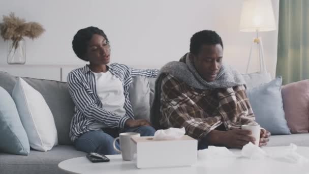 Malato ragazzo afroamericano che soffre di influenza a casa, seduto avvolto in plaid e bere il tè, sua moglie lo abbraccia — Video Stock