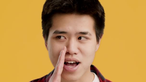 Азиатский парень шепчет, делясь секретами на жёлтом фоне — стоковое видео