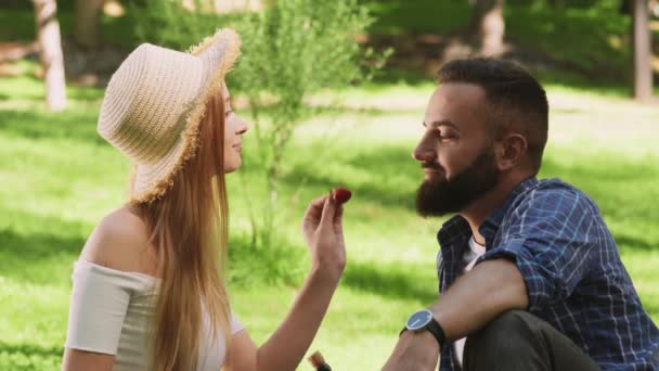 Gry miłosne. Uczuciowa para odpoczywająca w parku, karmiąca mężczyznę świeżą truskawką i całująca go — Wideo stockowe