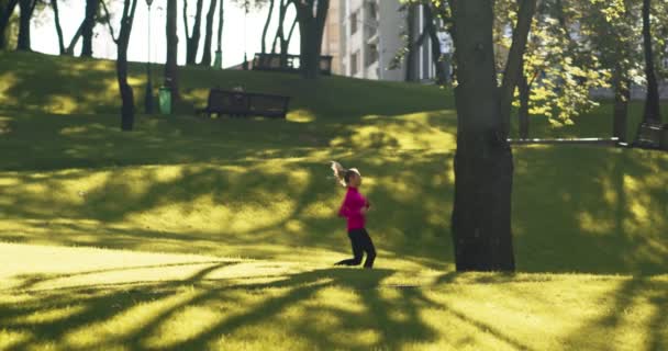 Jovem senhora loira desportiva em casaco rosa brilhante correndo sozinho no parque urbano vazio, praticando treino de corrida matinal — Vídeo de Stock