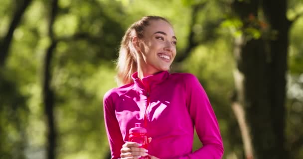 Conceito de estilo de vida saudável. Retrato de jovem senhora loira feliz descansando após o treino de corrida com garrafa de água no parque verde — Vídeo de Stock