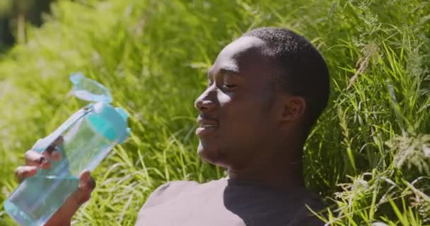 Koncepcja bilansu wody. Młody, spragniony Afrykańczyk pijący słodką wodę z plastikowej butelki, leżący na trawie — Wideo stockowe