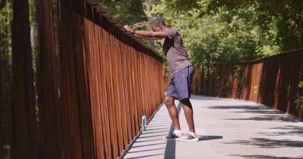 Sportig afrikansk amerikansk kille vilar på bron i stadsparken, står vid metall barer med vatten på morgonen, slow motion — Stockvideo