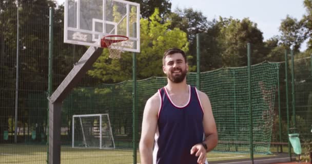Молодой позитивный бородатый спортсмен тренируется на открытом воздухе, играет в баскетбол, бросает мяч к обручу на площадке для стритбола — стоковое видео