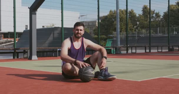 Außenporträt eines jungen aktiven Basketballspielers, der auf dem Boden des Tennisplatzes sitzt und mit Ball spielt und in die Kamera blickt — Stockvideo