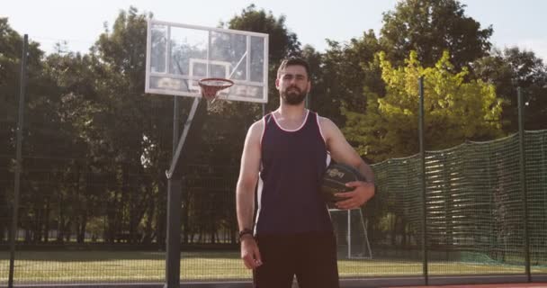 Kinematografický záběr mladého kavkazského basketbalisty pózujícího s míčem na venkovním hřišti streetballu, dívajícího se na kameru — Stock video