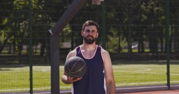 Спортивний бородатий білий баскетболіст в уніформі позує з м'ячем на відкритому майданчику, тренується сам — стокове відео
