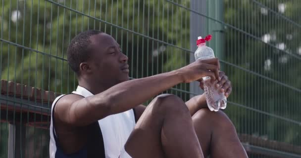 Νεαρός Αφροαμερικάνος ποδοσφαιριστής που κάνει διάλειμμα στην υπαίθρια παιδική χαρά, κάθεται και πίνει νερό — Αρχείο Βίντεο