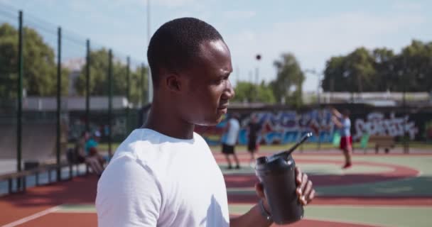 Mladý africký Američan pití vody ze sportovní láhve na venkovním basketbalovém hřišti, boční pohled, zpomalení — Stock video