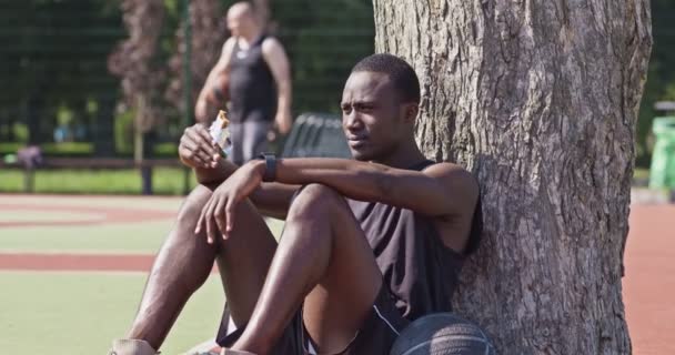 Молодой африканский баскетболист ест протеиновый бар, отдыхает после тренировки на площадке для стритбола, склоняется к дереву — стоковое видео