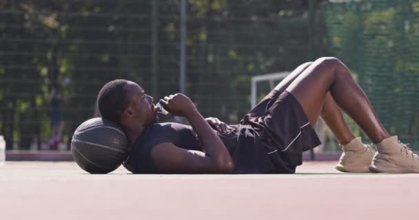 Νεαρός κουρασμένος Αφροαμερικάνος που αναπαύεται στο πάτωμα της παιδικής χαράς του δρόμου, ξαπλωμένος στο μπάσκετ και τρώγοντας μπάρες πρωτεΐνης. — Αρχείο Βίντεο