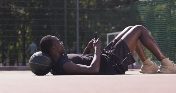 Joven jugador de baloncesto afroamericano en red en el teléfono inteligente, viendo el juego, descansando en la pelota, tumbado piso — Vídeos de Stock