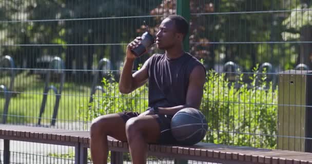 Giovane sudato sportivo afroamericano che riposa su panchina e acqua potabile, prendendo pausa nella partita di streetball — Video Stock