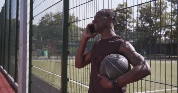 Θετικός Αφροαμερικάνος αθλητής μιλάει στο κινητό τηλέφωνο, περιμένοντας φίλους με μπάλα στο υπαίθριο γήπεδο μπάσκετ — Αρχείο Βίντεο