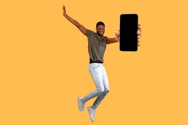 Крутий мобільний додаток. Захоплений чорний хлопець показує смартфон з порожнім чорним і стрибає на жовтому студійному фоні — стокове фото
