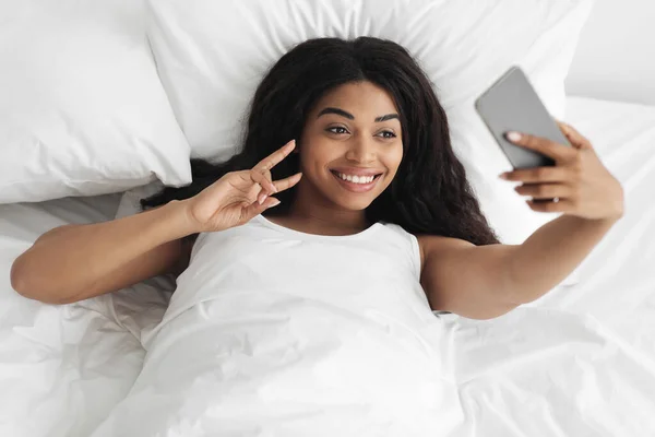 Счастливая черная женщина делает селфи на смартфоне, лежа утром в постели, фотографируя для социальных сетей или друзей — стоковое фото