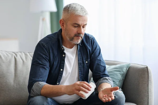 Senior man taking pills while sitting on couch at home — Stock Photo, Image