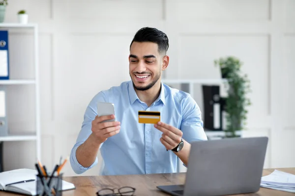 Zakupy online. Szczęśliwy arabski biznesmen posiadający kartę kredytową i smartfona, dokonujący płatności elektronicznej, zamawiający towary w internecie — Zdjęcie stockowe