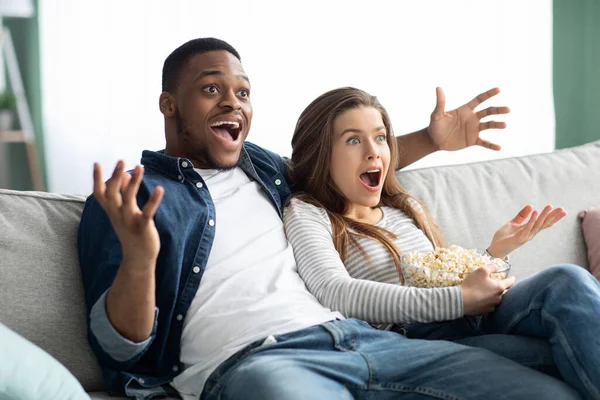 Emocionada pareja interracial viendo la televisión en el sofá en casa —  Fotos de Stock