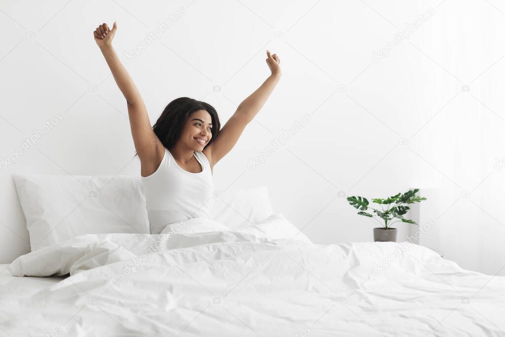 Relaxed after good night sleep. Happy african american woman sitting on bed with blanket, stretching after waking up