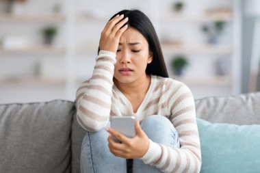 Endişeli Asyalı kadın akıllı telefon ekranına bakıyor, evdeki koltukta oturuyor.