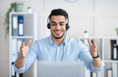 Telekomünikasyon kavramı. Kulaklıklı mutlu bir Arap dizüstü bilgisayarda çalışıyor, ofiste video görüşmesi yapıyor.