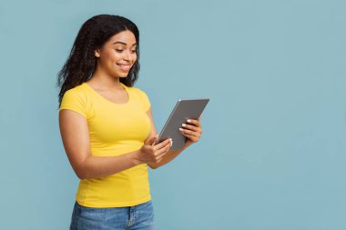 Mutlu Afro-Amerikalı kadın dijital tablet üzerinde sosyal ağlara göz atıyor, mavi stüdyo arka planında duruyor, boşluğu kopyalıyor.