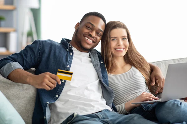 Pagos en línea. Retrato de cónyuges interraciales alegres con computadora portátil y tarjeta de crédito — Foto de Stock