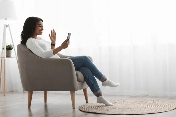 Africano senhora americana usando celular em casa, tendo videochamada on-line enquanto sentado em poltrona na frente da janela — Fotografia de Stock