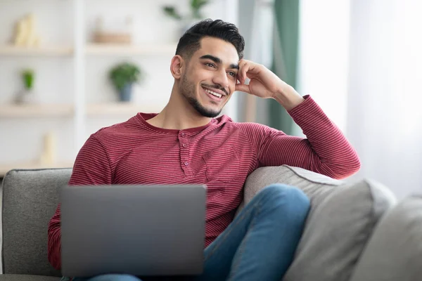 Dreamy arabski facet siedzi na kanapie z laptopem — Zdjęcie stockowe