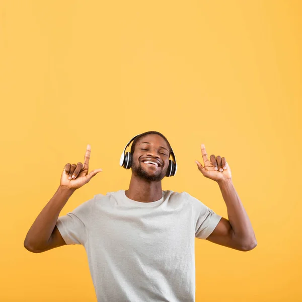 Захоплений чорний хлопець слухає музику в навушниках і піднімає пальці на вільний простір, жовтий фон, врожай — стокове фото