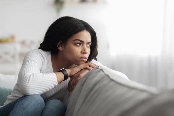 Concepto de depresión. Triste mujer negra sentada en el sofá y mirando a un lado en el espacio libre, sintiéndose sola —  Fotos de Stock