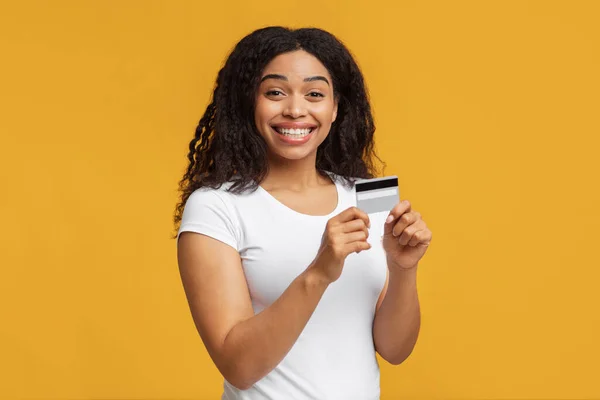 Łatwa cyfrowa płatność bezgotówkowa. Szczęśliwa czarna kobieta trzyma kartę kredytową i uśmiecha się do kamery, żółty tło studio — Zdjęcie stockowe