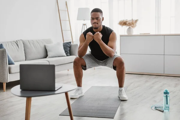 Sportowy Afrykanin robi głębokie przysiady w pobliżu laptopa komputer wewnątrz — Zdjęcie stockowe