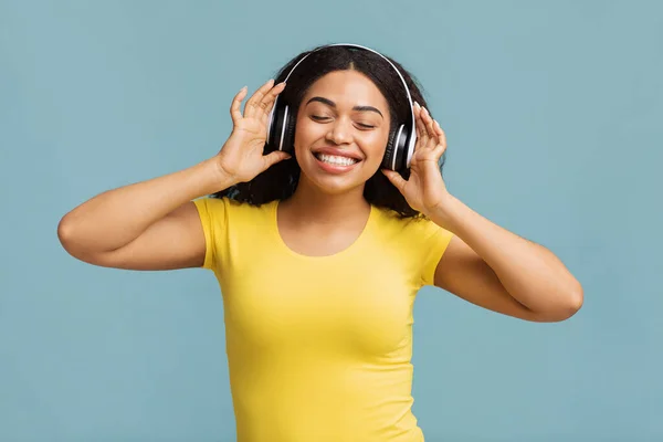Femme noire réjouie dans un casque sans fil écoutant de la musique avec les yeux fermés, fond bleu studio — Photo