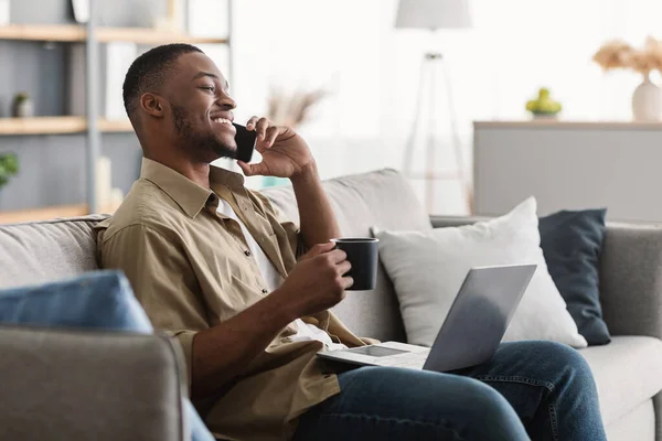 Hombre negro hablando en el teléfono celular usando el ordenador portátil beber café en interiores —  Fotos de Stock