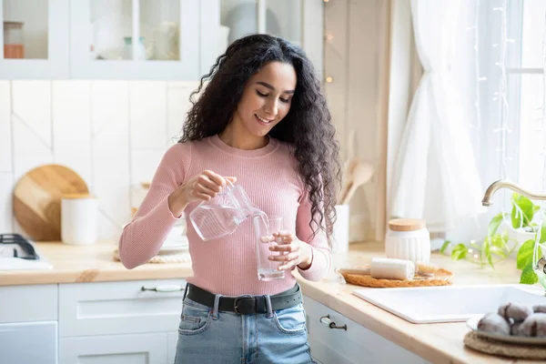 Hermosa joven dama vertiendo agua de jarra a vaso en la cocina —  Fotos de Stock