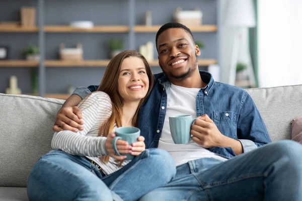 Домашнє дозвілля. Весела міжрасова пара дивиться телевізор і п'є каву на дивані — стокове фото