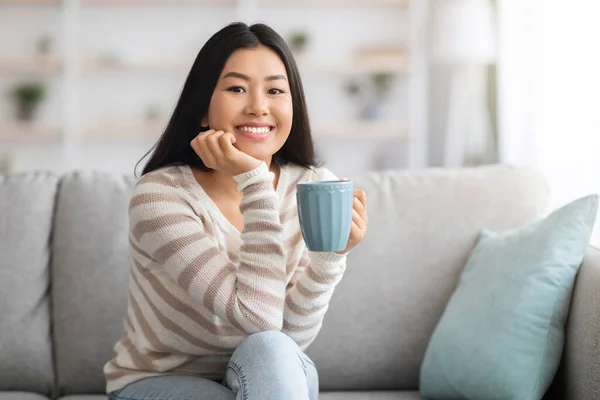 Домашний отдых. Счастливая улыбающаяся азиатка пьет кофе на диване дома — стоковое фото