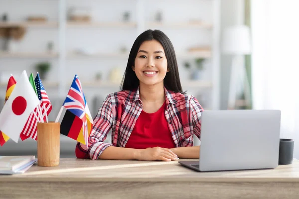 Glimlachende aziatische dame zitten aan tafel met internationale vlaggen en laptop — Stockfoto