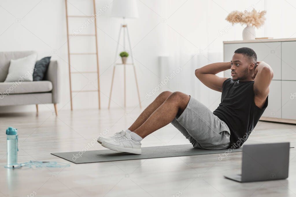 Side-View Of African American Guy Doing Sit-Ups At Laptop Indoor