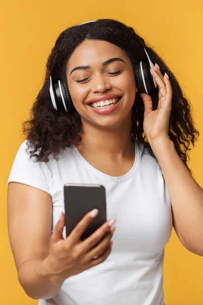 Boldog fekete hölgy élvezi a legújabb sztereó headset és mobil zenei alkalmazás, zenét hallgatni sárga stúdió háttér — Stock Fotó