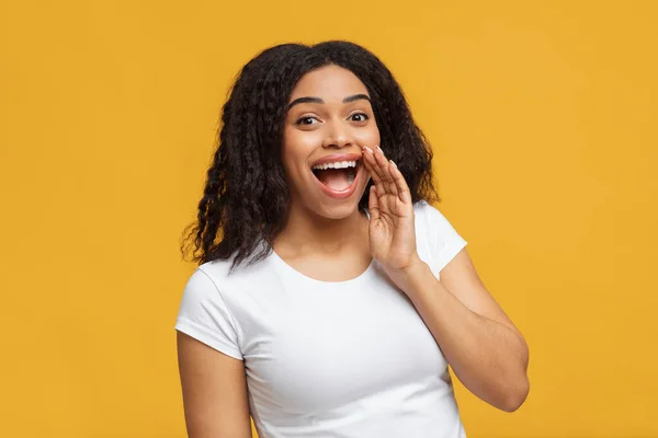 Femme afro-américaine partage des nouvelles, tenant la main près de la bouche ouverte et révélant le secret sur fond de studio jaune — Photo