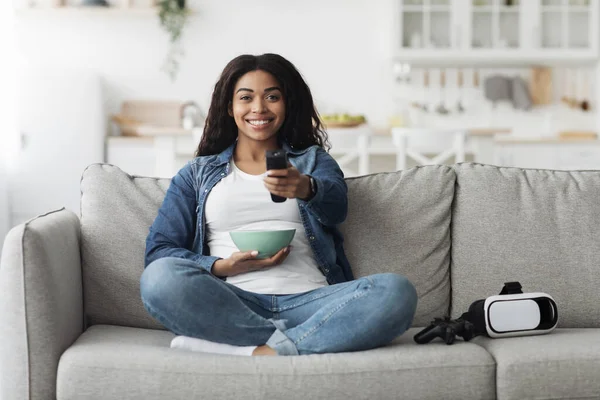 Entretenimiento en casa. Feliz dama negra con control remoto de búsqueda de película en la televisión, sentado en el sofá con gafas VR cerca —  Fotos de Stock