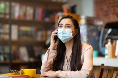 Arap kadın yüz maskesi takıyor ve akıllı telefondan konuşuyor, kafenin içinde masada oturuyor, boş yer var.