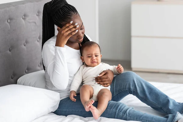 Уставшая афроамериканская молодая мама сидит с ребенком на кровати — стоковое фото