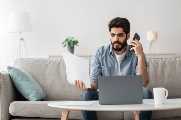 Problemas no trabalho. Homem árabe discutindo documentos, segurando papel e falando no celular, trabalhando remotamente em casa — Fotografia de Stock